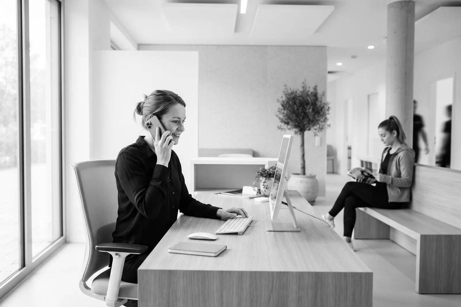 Receptionist on the phone at a modern clinic front desk while a patient waits